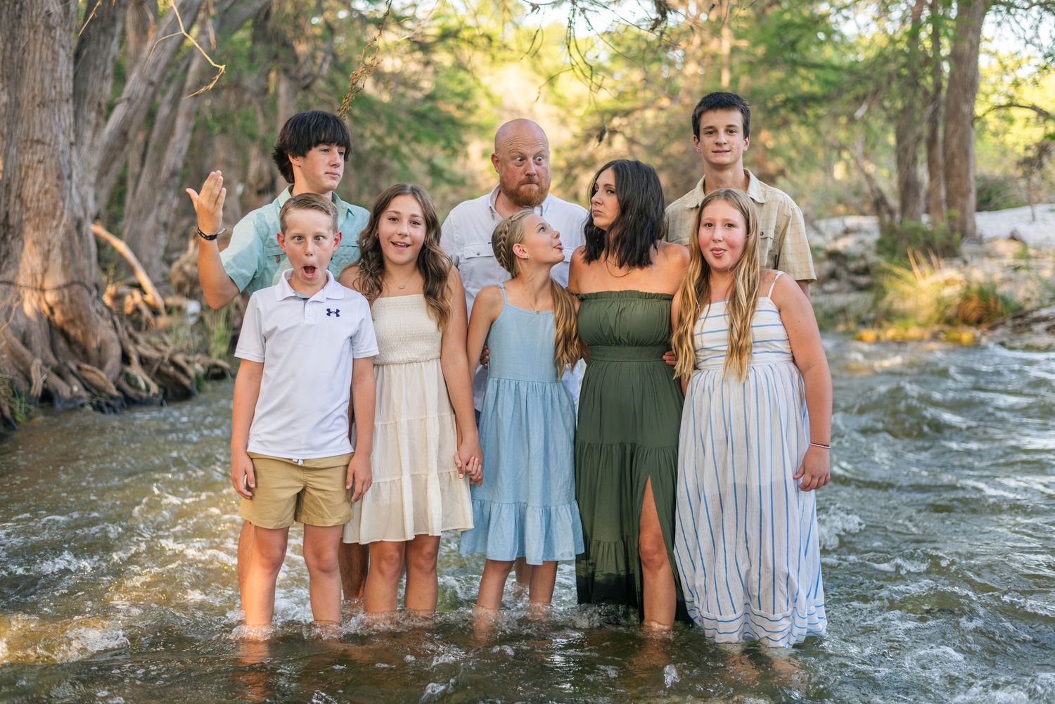 Family of eight posing together in a shallow river surrounded by trees during a outdoor photo session.