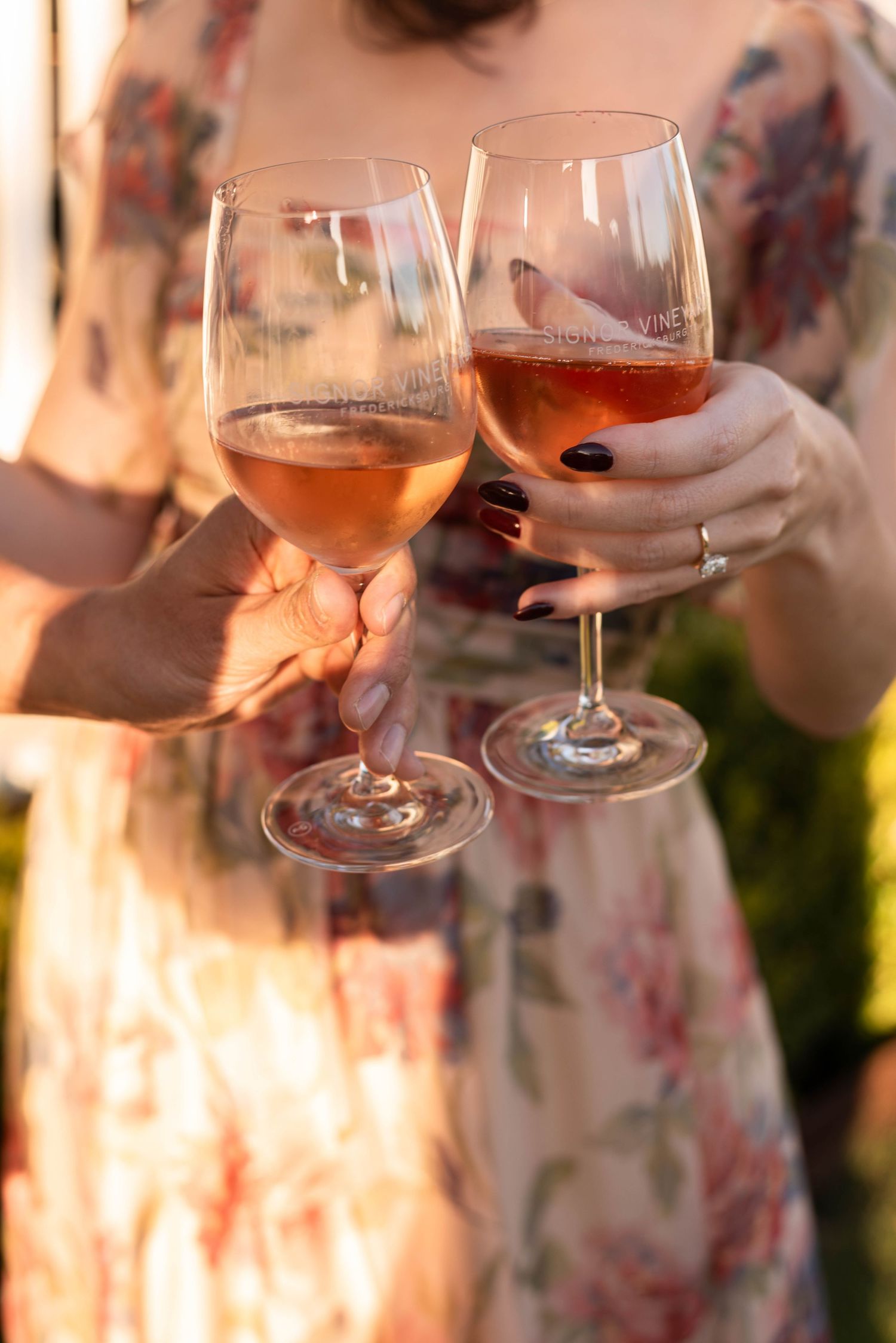 Two women toasting with glasses of rosé wine outdoors in golden sunset light, wearing floral dresses.
