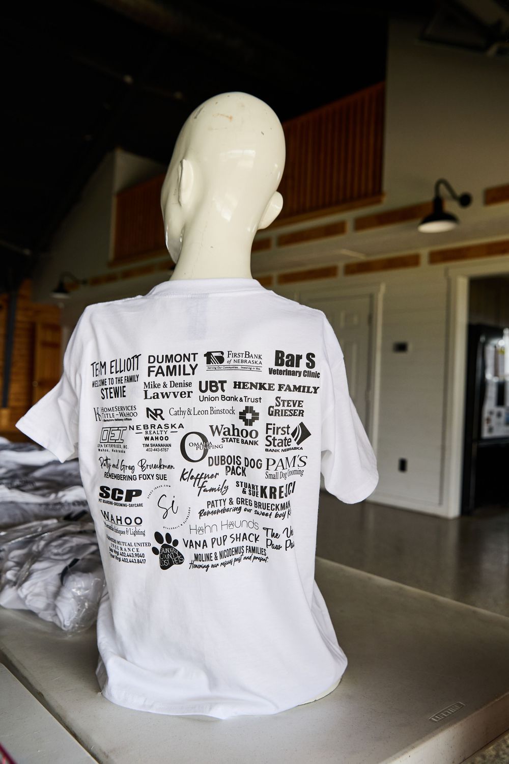 White sponsor t-shirt displayed on mannequin showing multiple business logos including Wahoo, UBT, and Henke Family.