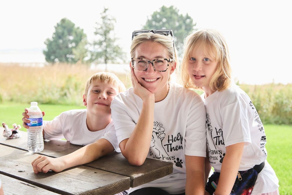 Three smiling children wearing matching white t-shirts pose together at a picnic table outdoors on a sunny day.