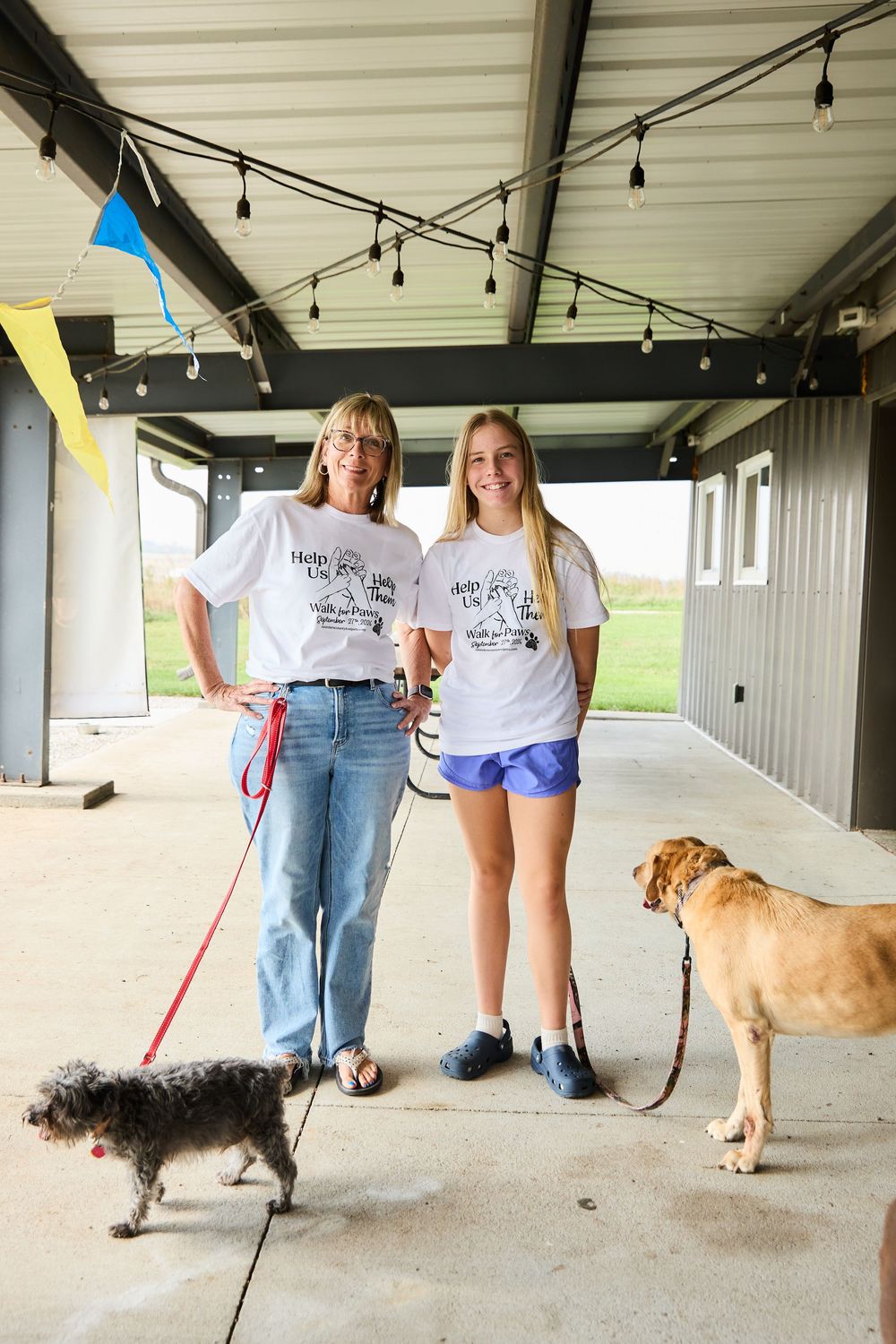 Two women in matching white t-shirts smile with two dogs on leashes inside a covered outdoor venue.