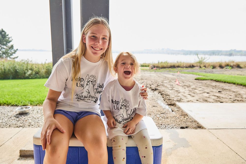 Two smiling girls in matching white t-shirts sit together on a cooler near a waterfront on a sunny day.