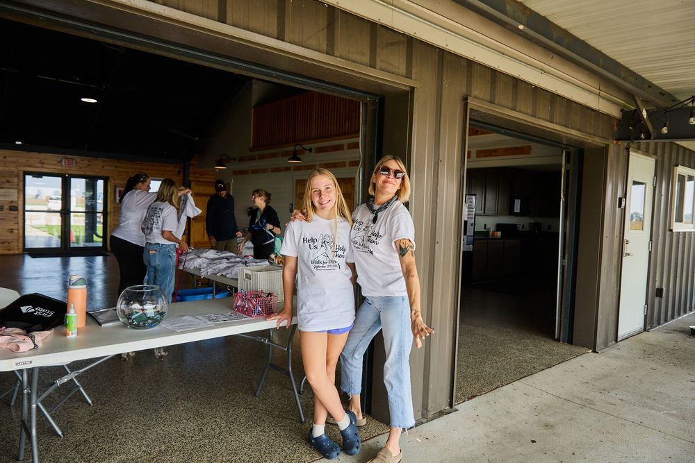 Two women in matching gray t-shirts pose together at an outdoor community event with tables and volunteers in background.