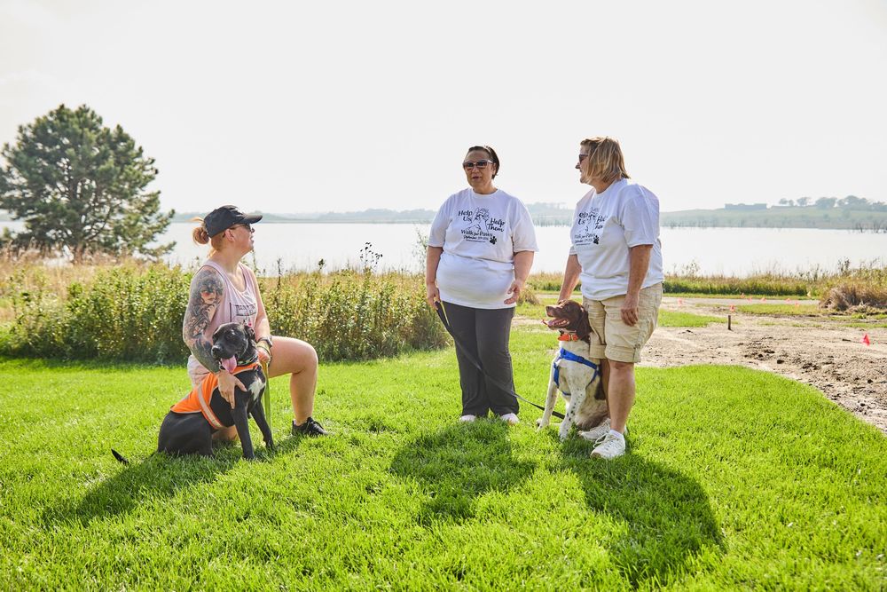 Three women with dogs gather on green grass near a lake on a sunny day, wearing matching white t-shirts.