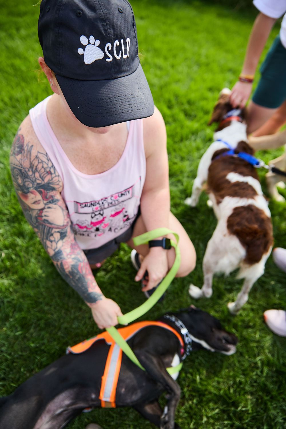 Tattooed woman in SCLP cap holds leashes of two dogs on green grass at outdoor pet event.