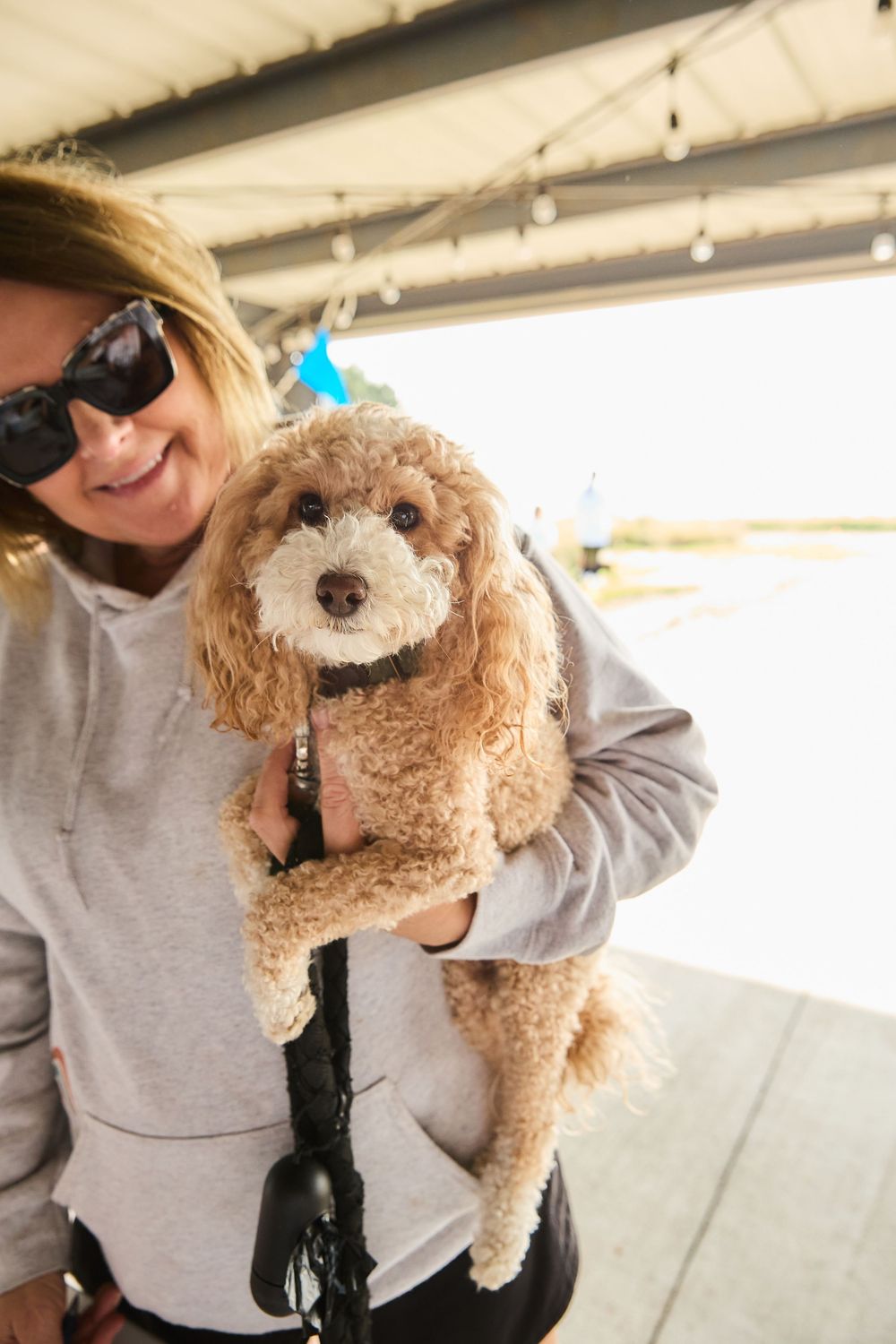 Smiling woman in gray hoodie and sunglasses holding a fluffy brown poodle mix dog outdoors.