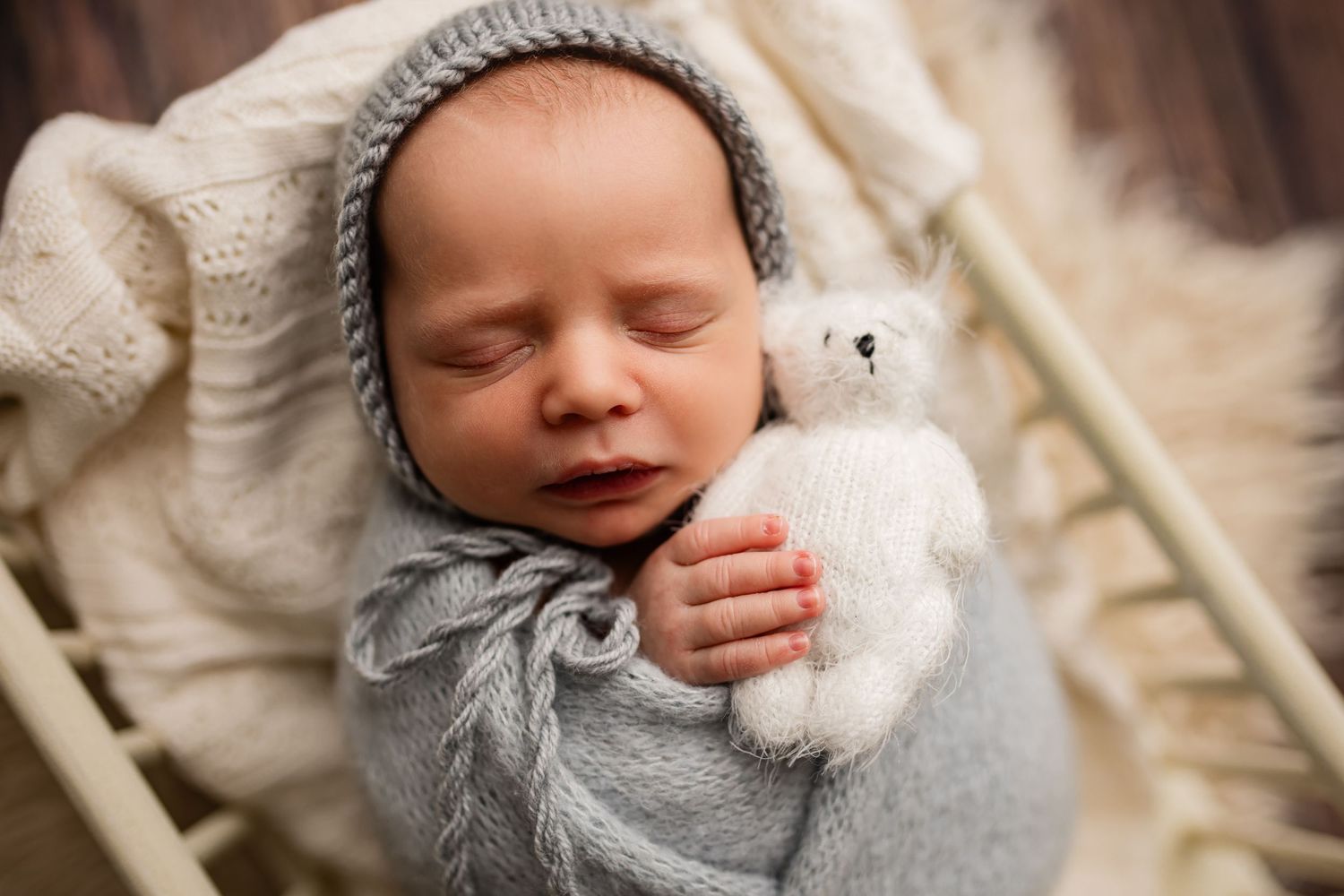 Sleeping newborn baby wrapped in gray knit blanket wearing bonnet holding small white teddy bear in cozy basket.