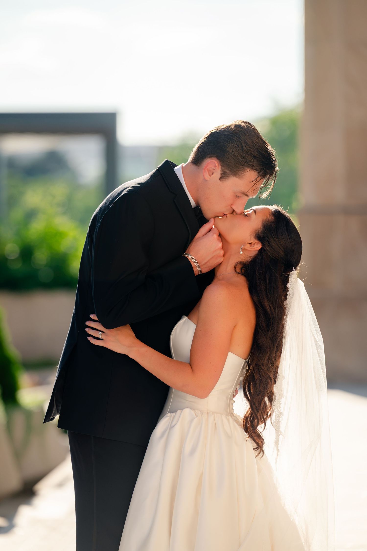 A bride and groom share a romantic kiss outdoors, with the bride in a strapless white gown and veil.