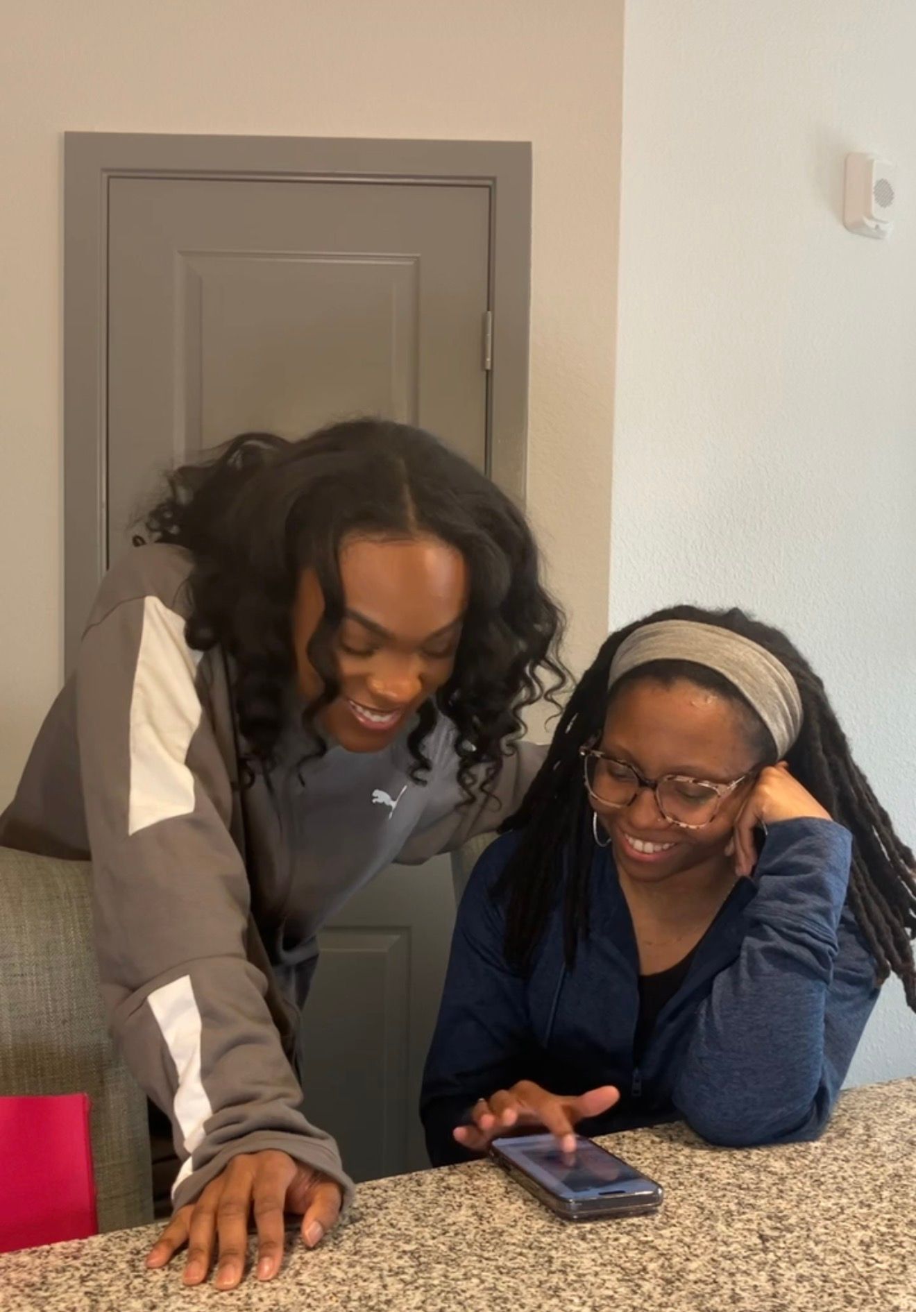 Two young women smiling and looking at a smartphone together indoors near a counter.