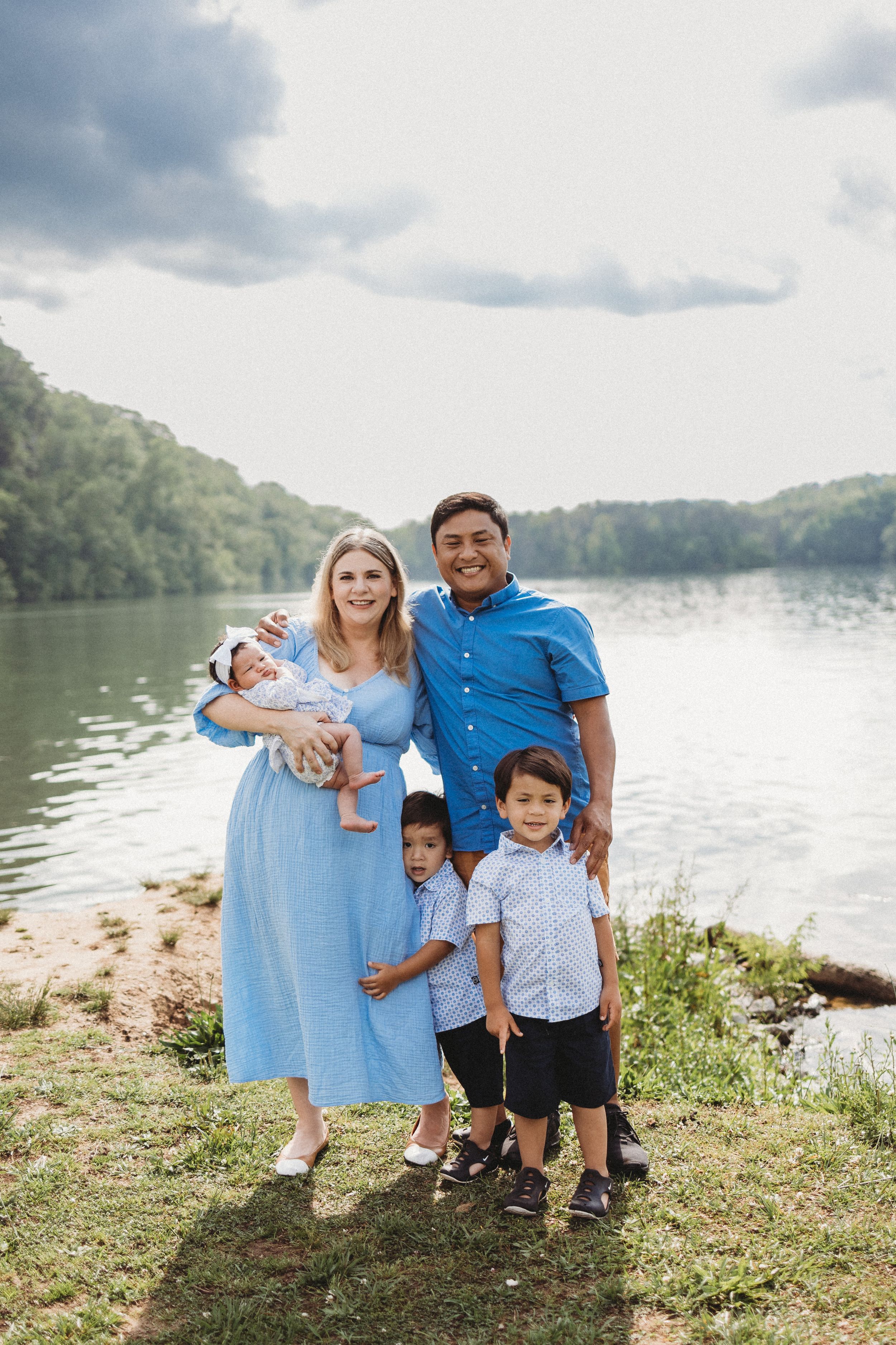 Family of five posing by a scenic lake, parents in blue outfits with three young children on a sunny day.