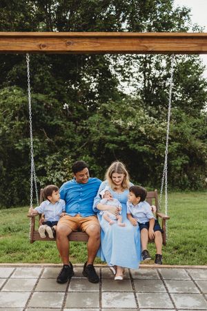 Family of five sitting on a wooden porch swing outdoors, parents holding newborn baby flanked by two young boys.
