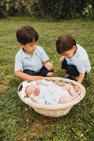 Two boys in blue shirts watch over a sleeping newborn in a wicker basket on green grass.