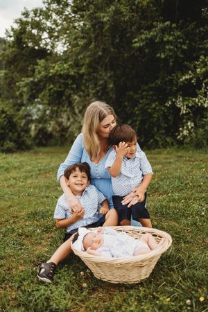A mother sits on grass with two young boys in matching blue shirts and a newborn baby in a wicker basket outdoors at Melton Hill Park
