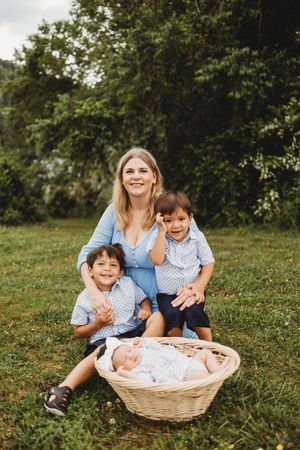 A smiling mother sits on grass with two young boys and a newborn baby in a wicker basket, outdoors at Melton Hill Park in Knoxville, TN