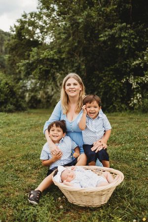 Smiling mother in blue dress sits on grass with two young boys and newborn baby in wicker basket outdoors at Melton Hill Park in Knoxville