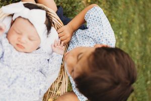 Older child lovingly gazes at sleeping newborn baby in a wicker basket outdoors in a garden setting at Melton Hill Park in Knoxville, TN