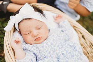 Newborn baby girl sleeping peacefully in a wicker basket, wearing a floral outfit and white bow headband outdoors at Melton Hill Park