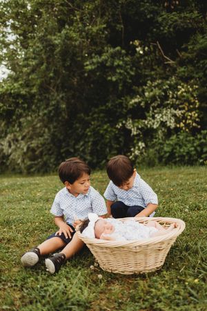 Two young brothers in matching blue shirts sit on grass beside wicker basket holding sleeping newborn baby at Melton Hill Park in Knoxville,
