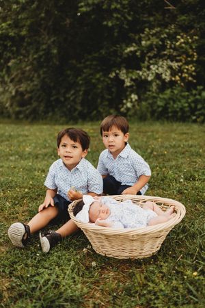 Two young boys in matching blue patterned shirts pose beside wicker basket with newborn baby on green grass at Melton Hill Park in Knoxville