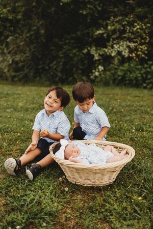 Two young brothers in matching blue shirts sit beside a wicker basket with a sleeping newborn outdoors at Melton Hill Park in Knoxville, TN