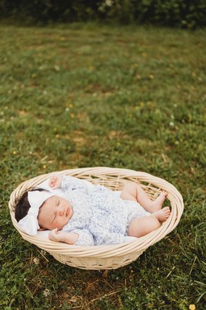 Sleeping newborn baby in blue floral outfit and white bow lying in a wicker basket on green grass outdoors at Melton Hill Park in Knoxville,