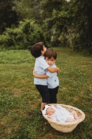 Two young boys hugging outdoors near a newborn baby sleeping in a wicker basket on green grass at Melton Hill Park in Knoxville, TN