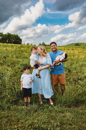Family of four poses in a sunny wildflower field, mom kissing toddler while dad holds newborn baby at Melton Hill Park in Knoxville, TN