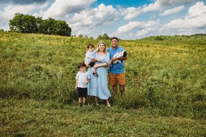 Family of four posing in a sunny green meadow with wildflowers, mother in blue dress holding newborn baby at Melton Hill Park in Knoxville,