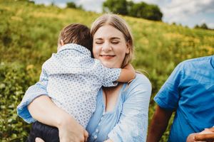 A smiling mother holds her young child in a blue shirt outdoors in a green field on a sunny day at Melton Hill Park in Knoxville, TN