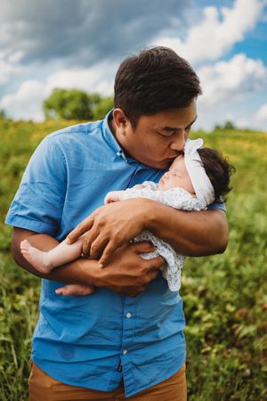 A father tenderly kisses his newborn baby while standing in a green field under a cloudy sky at Melton Hill Park in Knoxville, TN