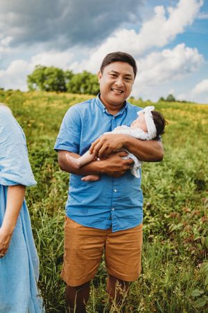 Smiling father in blue shirt holds newborn baby in a lush green wildflower meadow at Melton Hill Park in Knoxville, TN