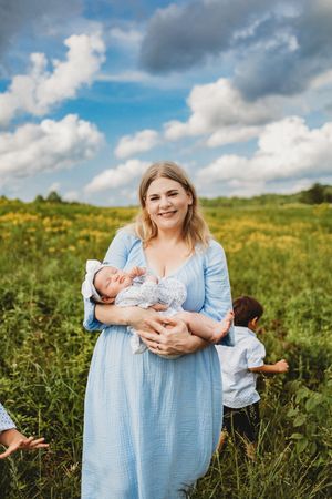 A smiling woman in a blue dress holds a newborn baby in a sunny green field with yellow wildflowers at Melton Hill Park in Knoxville, TN