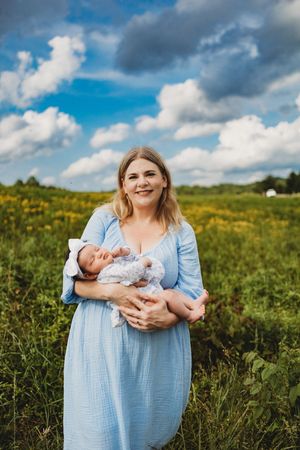 A smiling mother in a blue dress holds her newborn baby in a sunny green field with yellow wildflowers at Melton Hill Park in Knoxville, TN