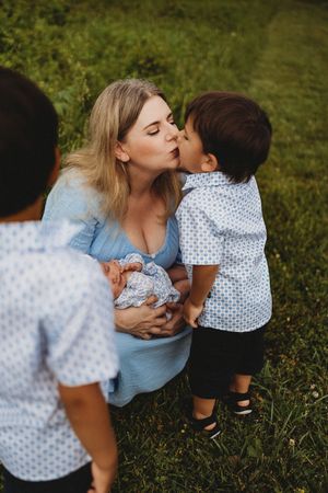 A blonde mother in a blue dress holds a newborn while a young boy gives her a kiss outdoors at Melton Hill Park in Knoxville, TN