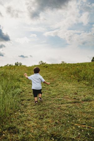 A young boy in a light blue shirt runs up a grassy hill under a cloudy sky at Melton Hill Park in Knoxville, TN