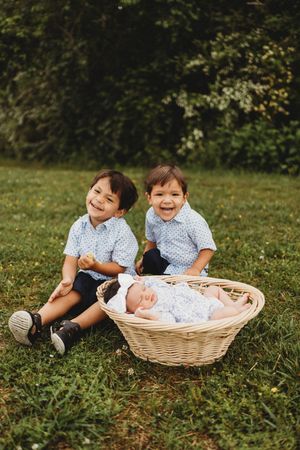 Two happy young boys in matching shirts sit on grass beside a wicker basket holding a sleeping newborn at Melton Hill Park in Knoxville, TN