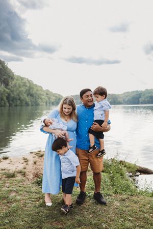 Happy family of five posing by a river, parents holding children in blue outfits on a cloudy day.