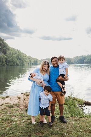 Family of five posing by a river, parents holding two toddlers, mother in blue dress cradling newborn baby.