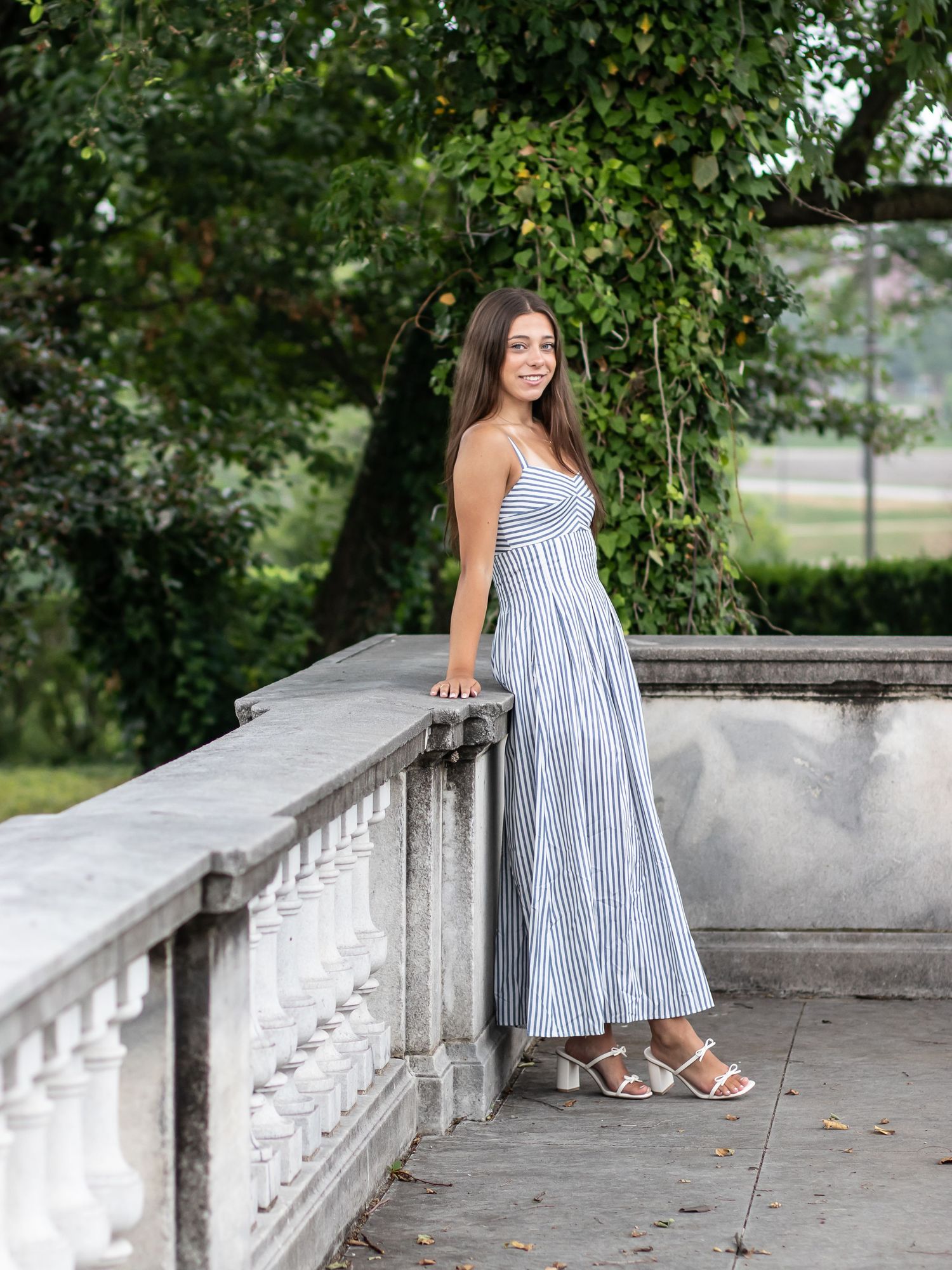 Young woman in striped sundress posing gracefully by stone balustrade surrounded by green ivy in outdoor setting.
