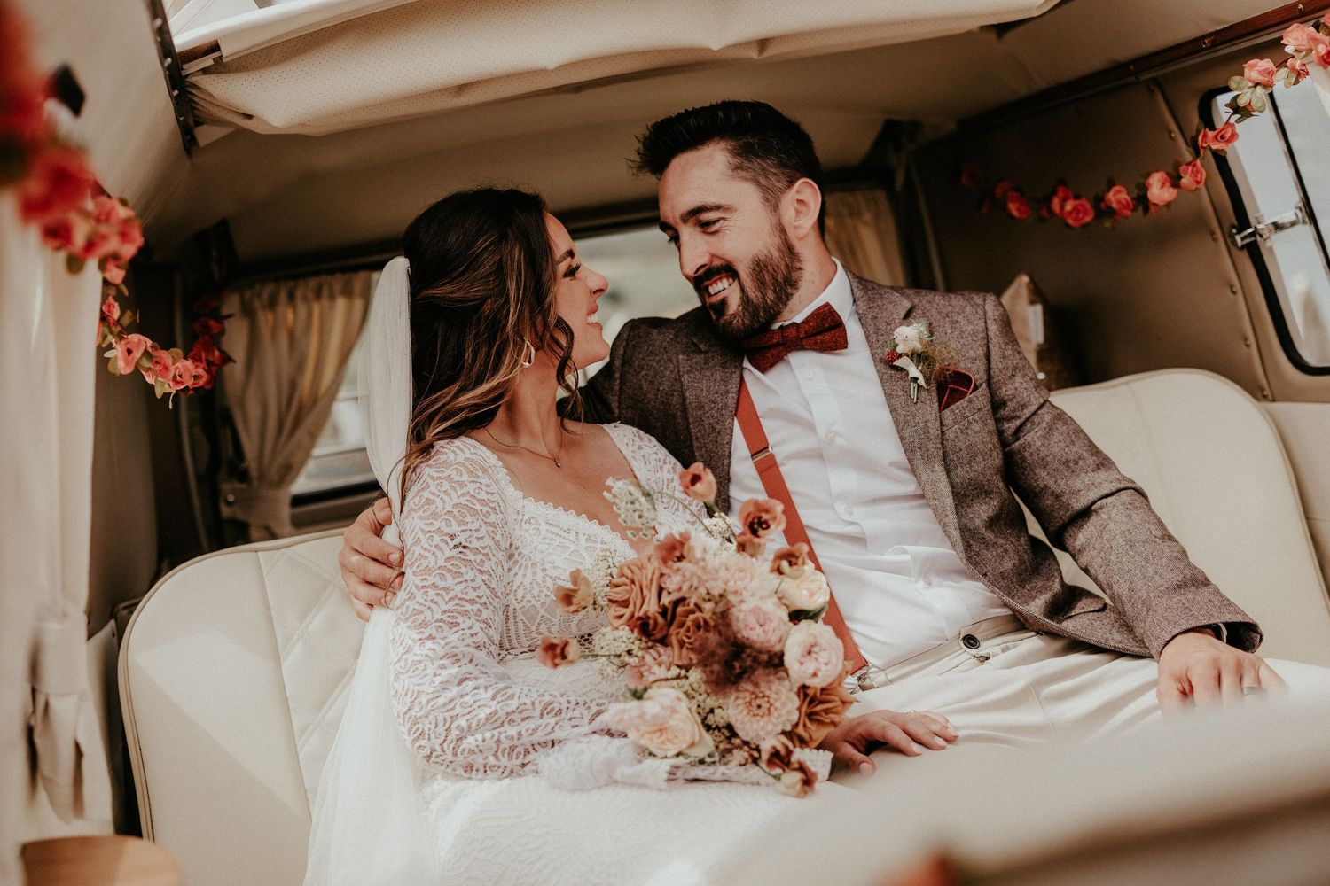 Bride and groom sitting together in a decorated vintage van, smiling at each other, bride holding a boho floral bouquet.