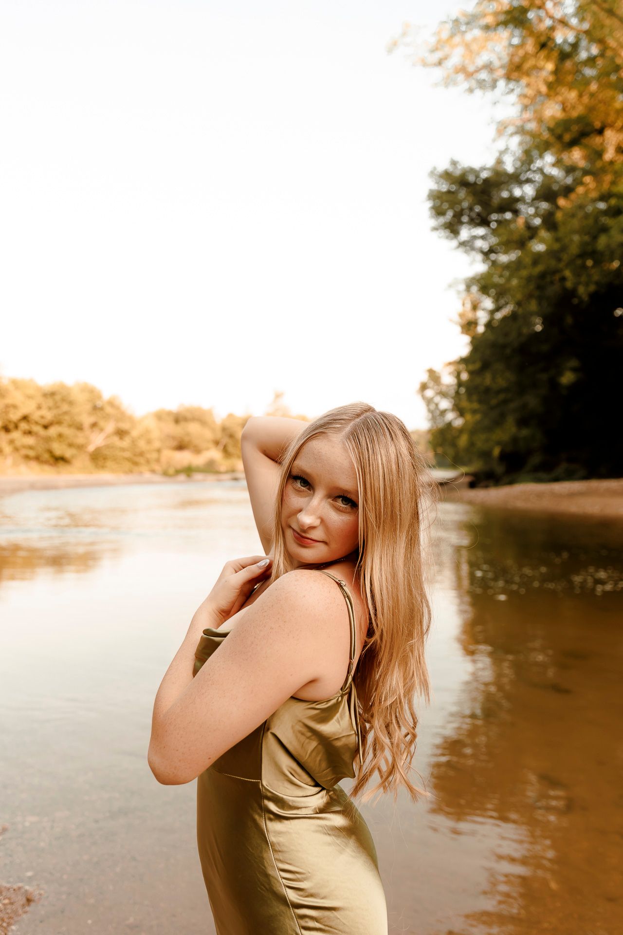 Young blonde woman in olive satin dress poses gracefully by a sunlit river with autumn trees in background.
