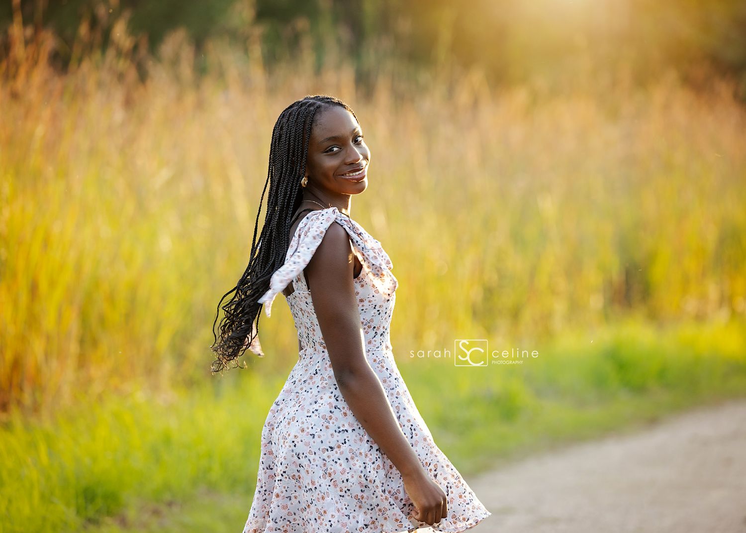 Young Black woman in floral dress poses on a dirt path surrounded by golden tall grass in warm sunlight.