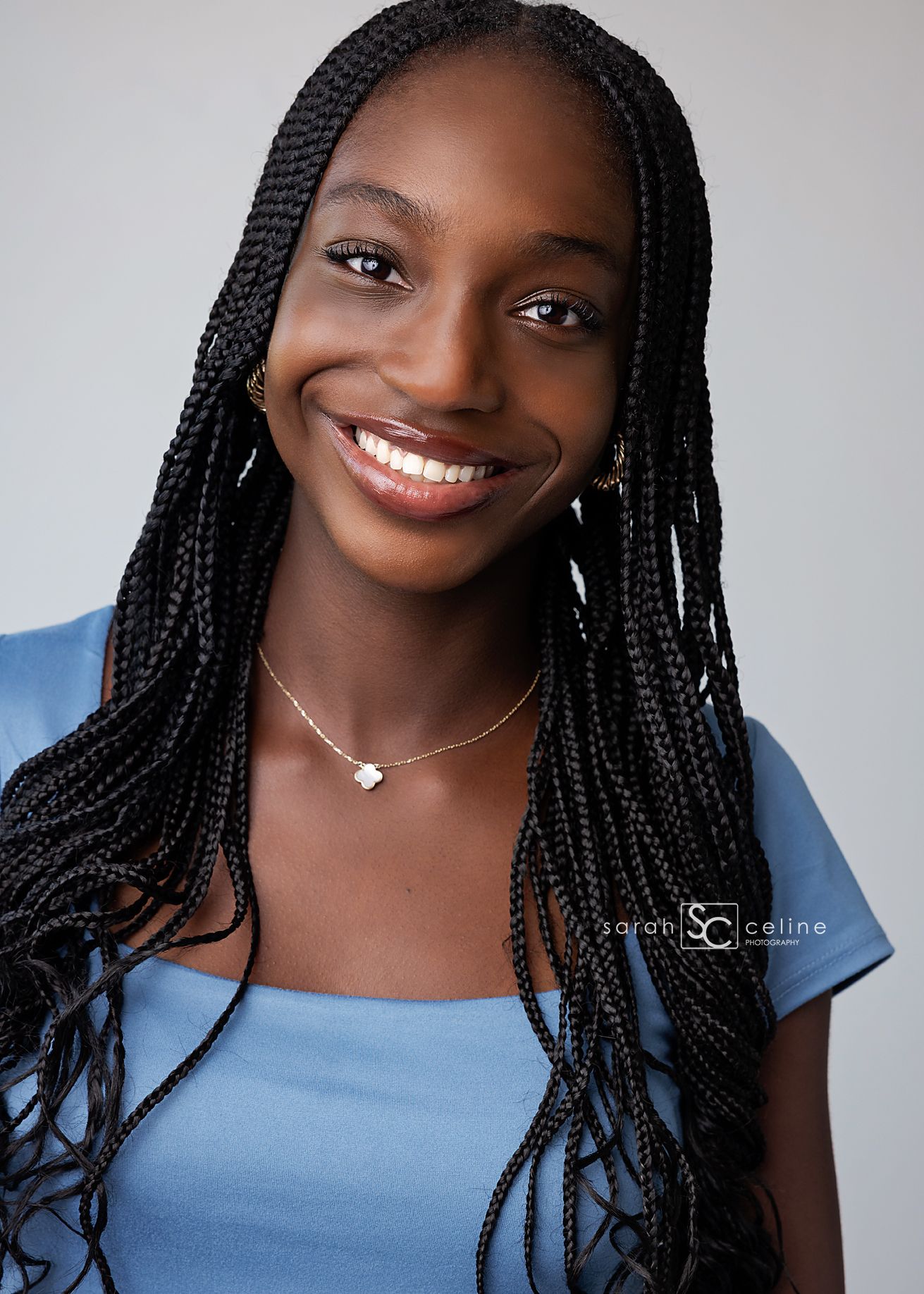 Young woman with long box braids smiling confidently in a blue top, wearing a delicate gold necklace.