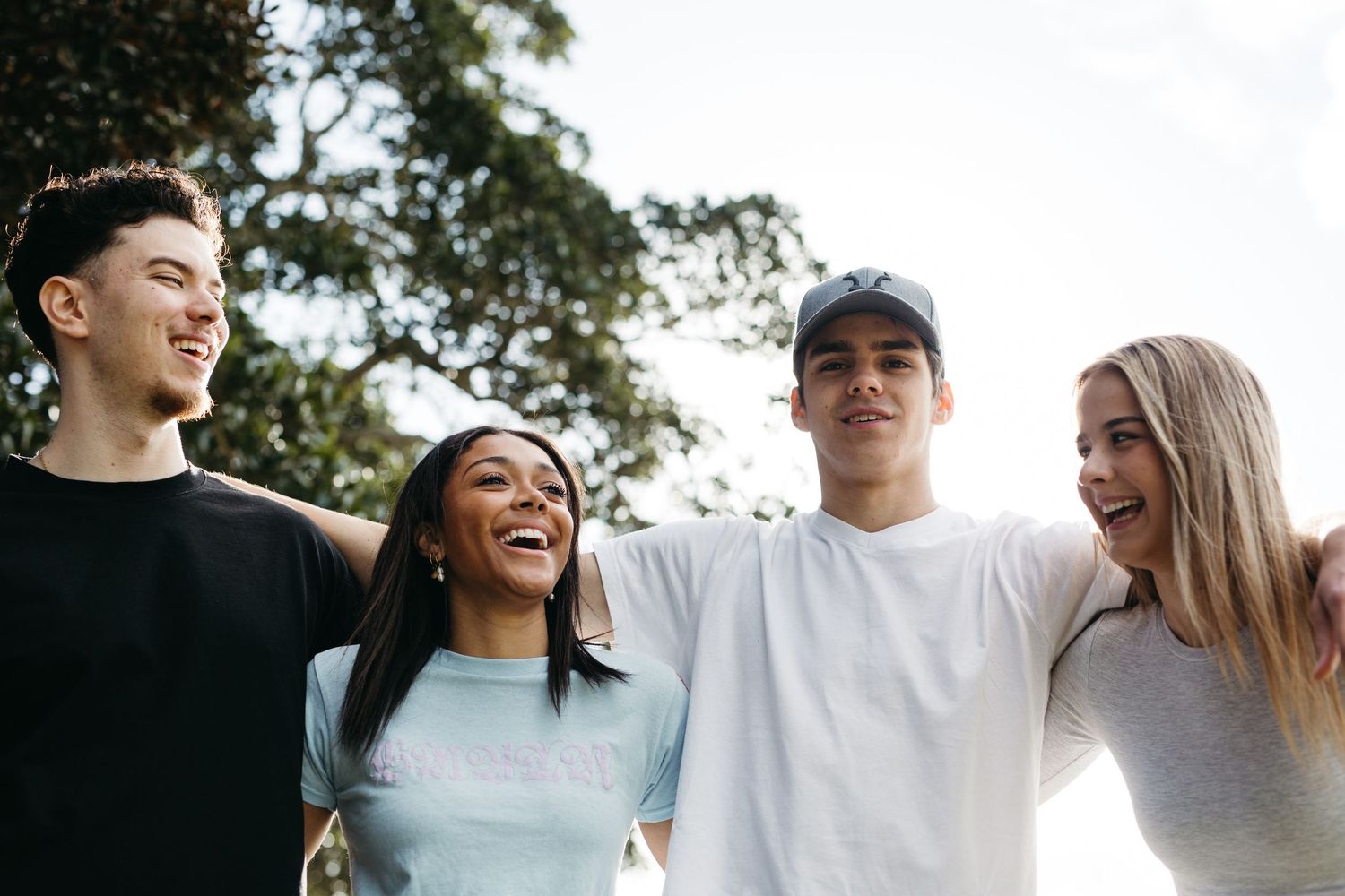 Four happy teenagers smiling and embracing outdoors in a park setting on a bright day.
