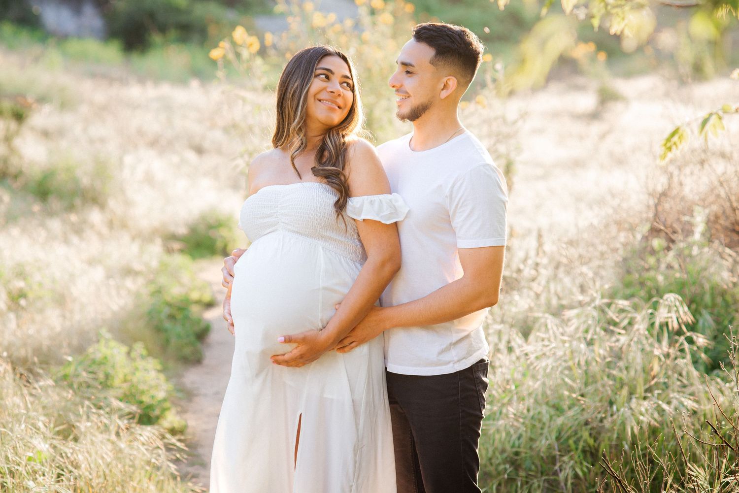 Couple in white embracing during maternity photoshoot in sunlit field.