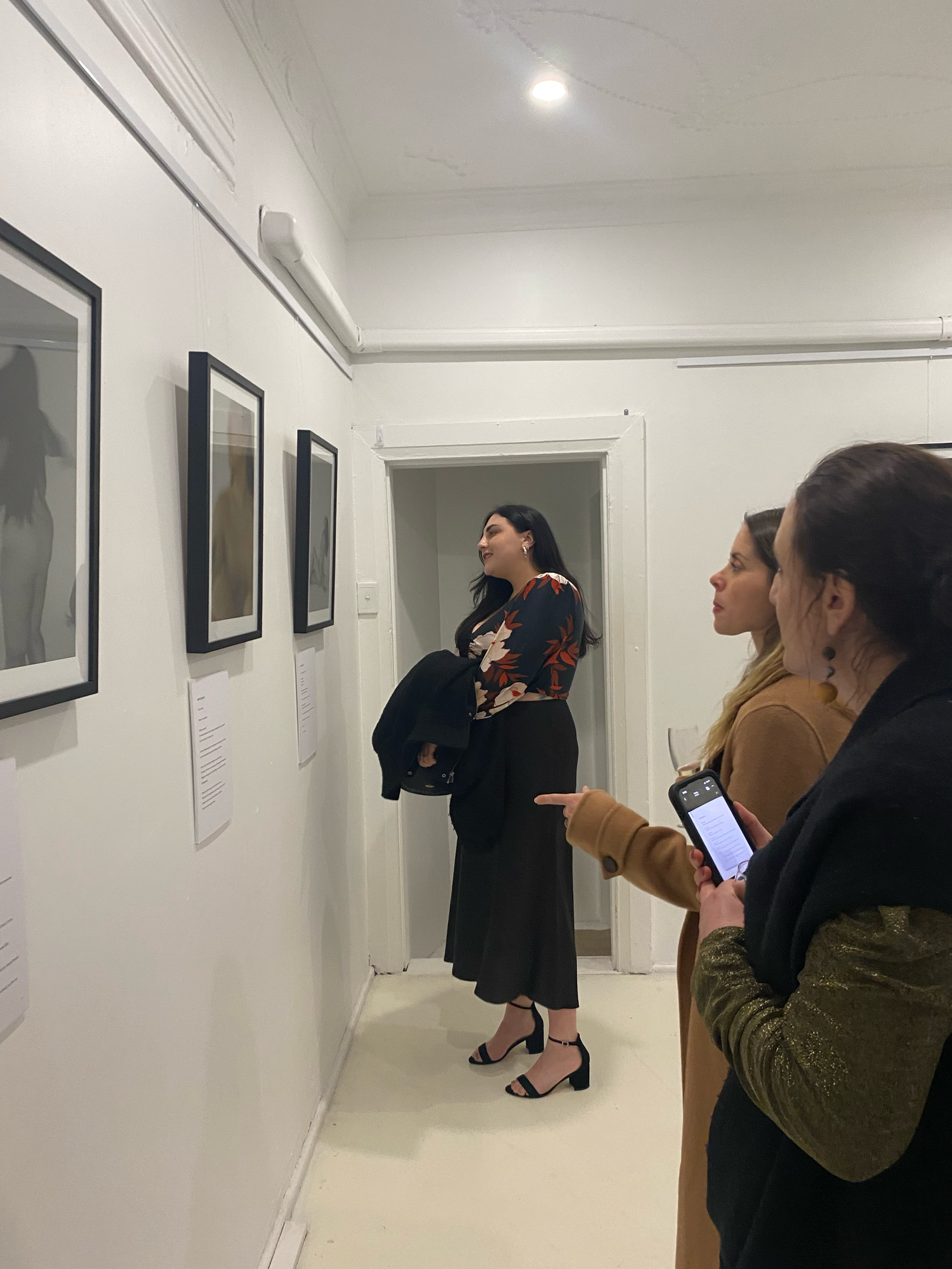 Three women viewing framed artwork on white gallery walls during an evening art exhibition.