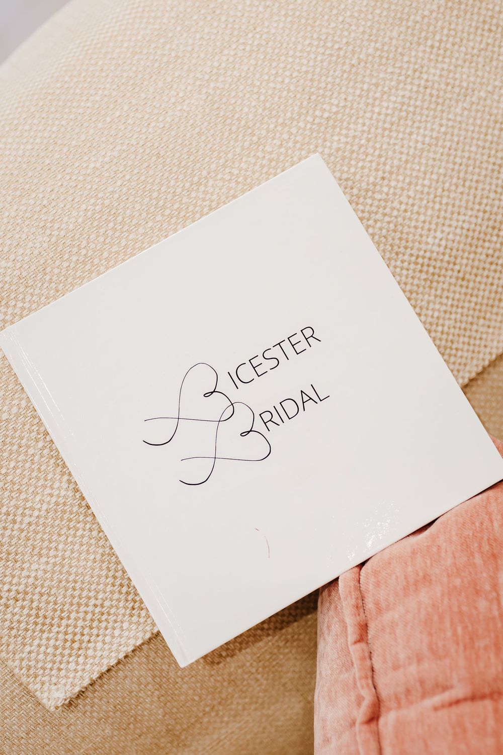 Bicester Bridal white logo book resting on a beige textured sofa with a pink cushion.