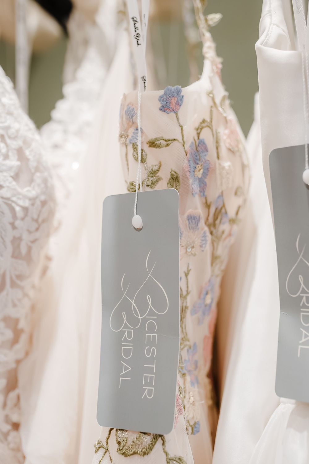Close-up of a floral embroidered wedding dress with a gray Bicester Bridal tag hanging in a bridal boutique.