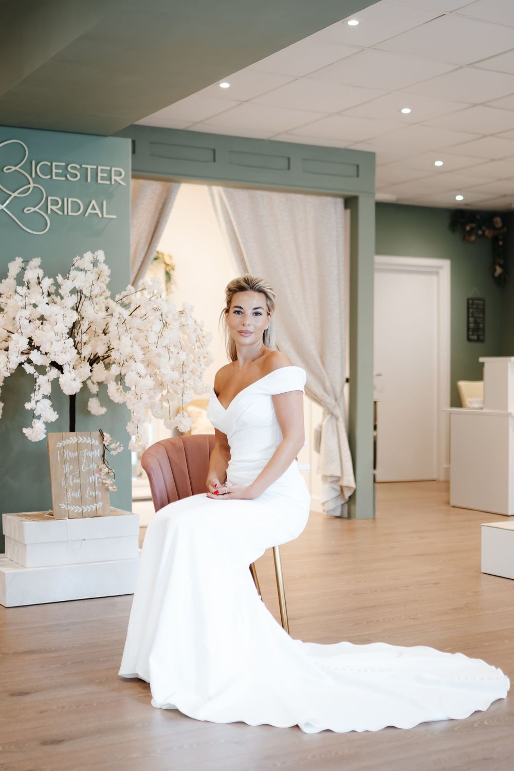 Bride in off-shoulder white gown seated elegantly at Bicester Bridal boutique with floral decor.