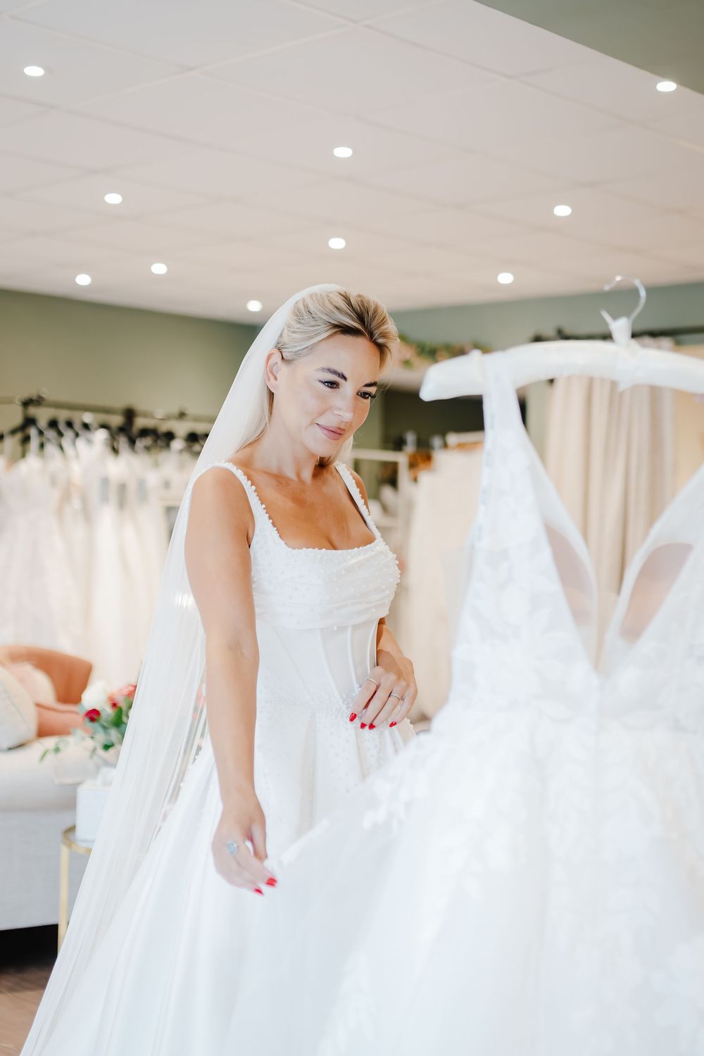 Blonde bride in white gown and veil admiring a lace wedding dress on a hanger in a bridal boutique.